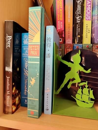 Four books next to a green bookstop featuring a silhouette of Peter Pan hovering above a distant pirate ship. From left to right, the books are: Peter: A Tale from Neverland by Jonathan M. Wenzel, Peter and the Starcatchers by Barry & Pearson, a Minalima illustrated version of Peter Pan, and a graphic novel version in Chinese. Behind the books, part of a dark blue ukulele is visible.